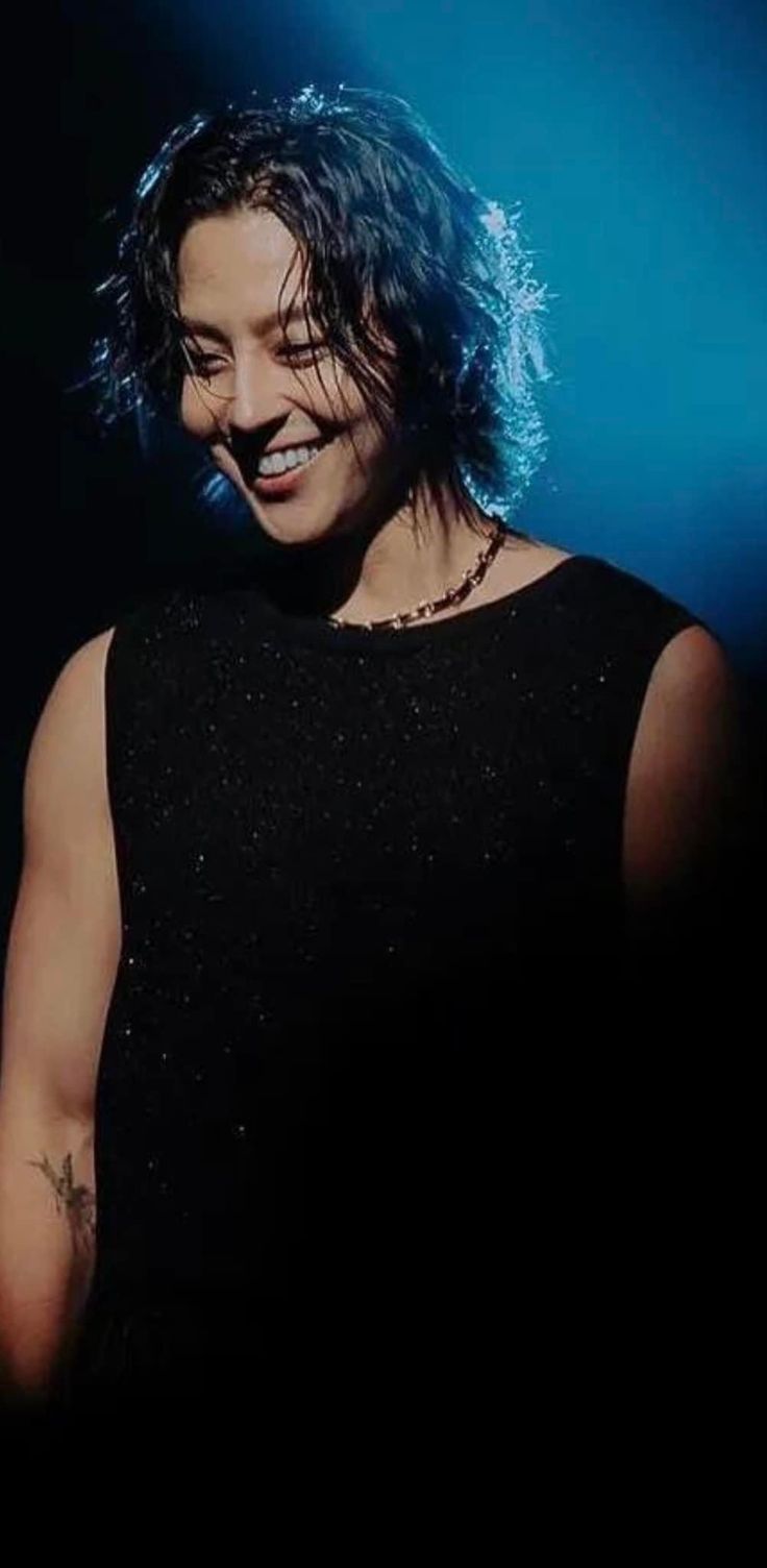 a woman with dark hair and piercings on her chest smiles at the camera while standing in front of a blue light