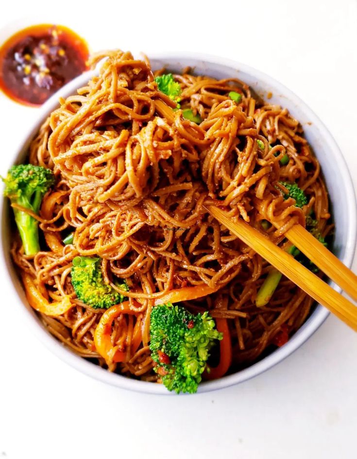 a bowl filled with noodles, broccoli and carrots next to some dipping sauce