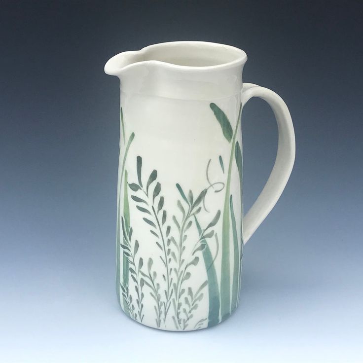 a white pitcher with green plants painted on the front and sides, sitting against a gray background