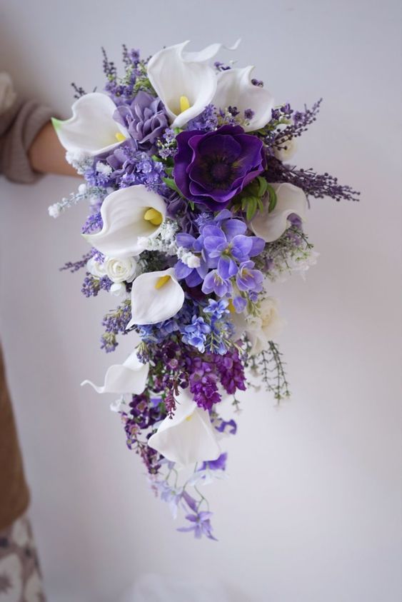 a bridal bouquet with purple and white flowers in the center is held up by someone's hand