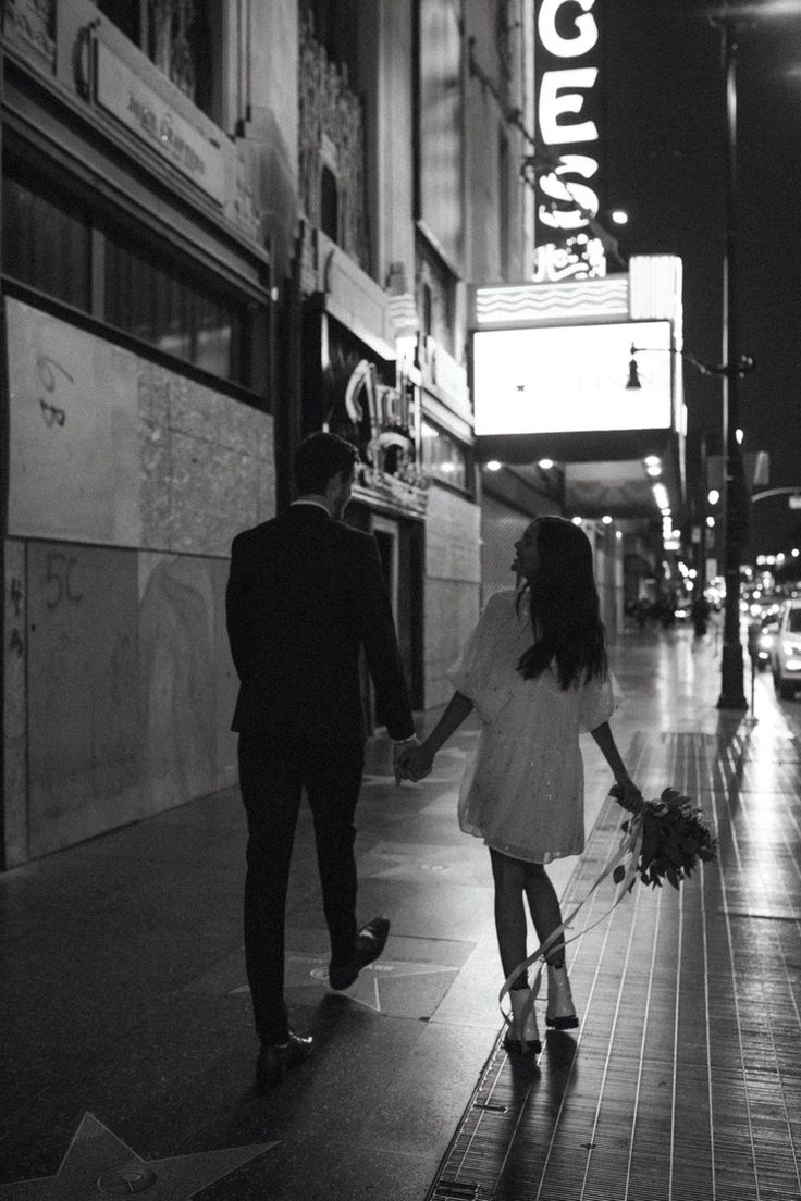 a man and woman holding hands walking down the street in front of a movie theater