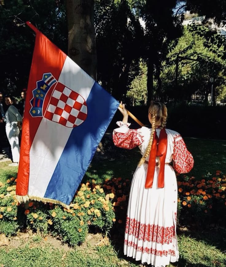 a woman in a long dress holding an croatia flag on the grass with other people behind her