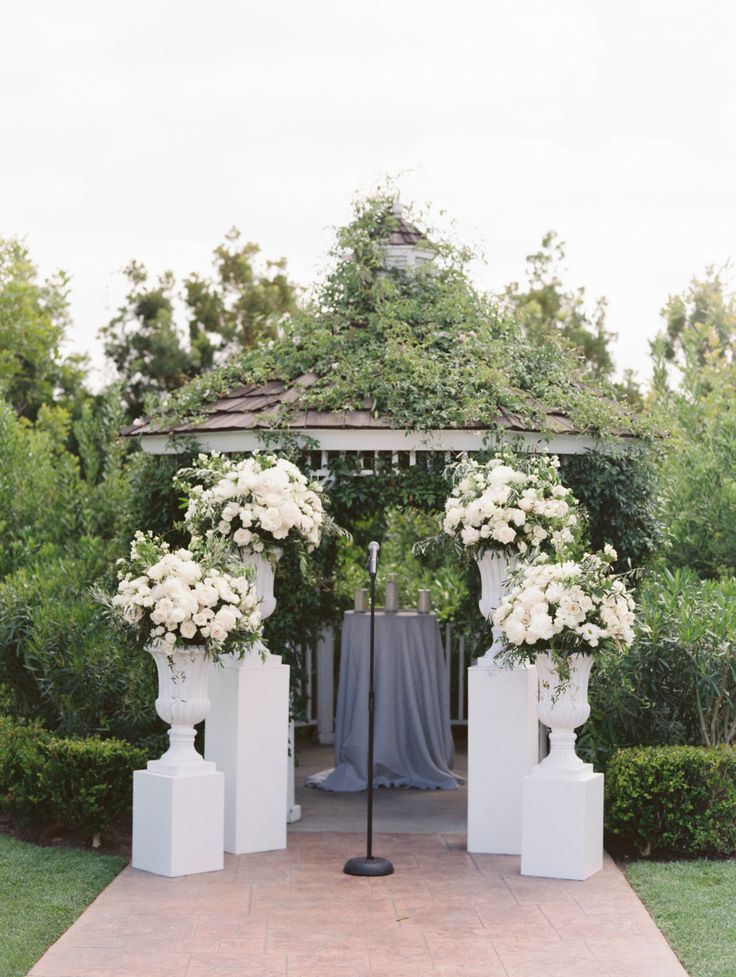 a gazebo with white flowers and greenery on the top is surrounded by bushes