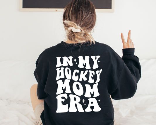 a woman sitting on a bed wearing a black hoodie that says in my hockey mom era