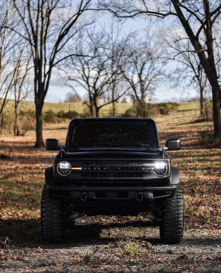 the front end of a black truck parked on top of a dirt road next to trees