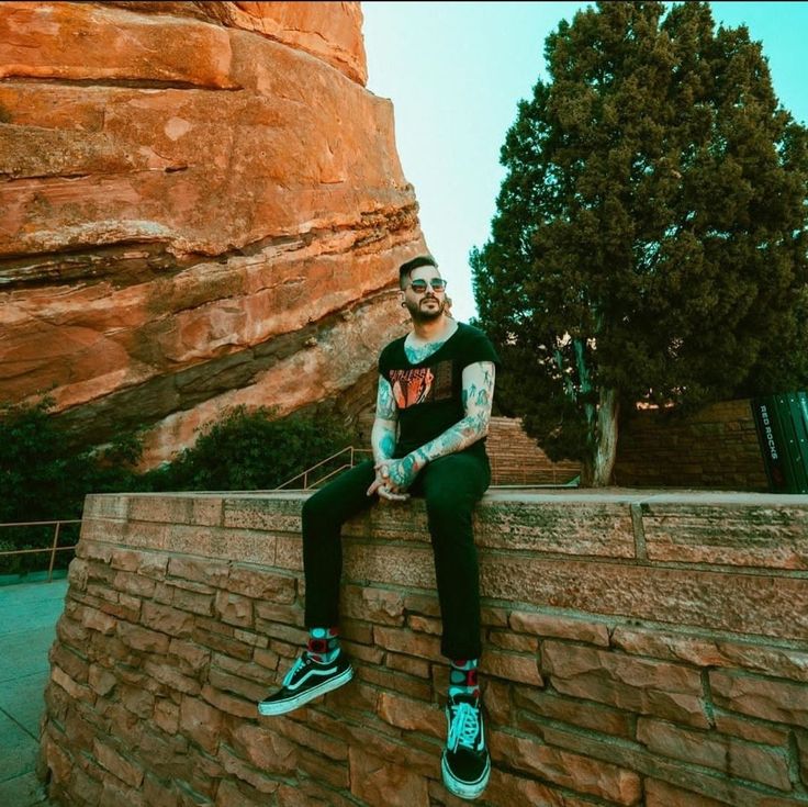 a man sitting on top of a brick wall next to a large rock formation with trees in the background