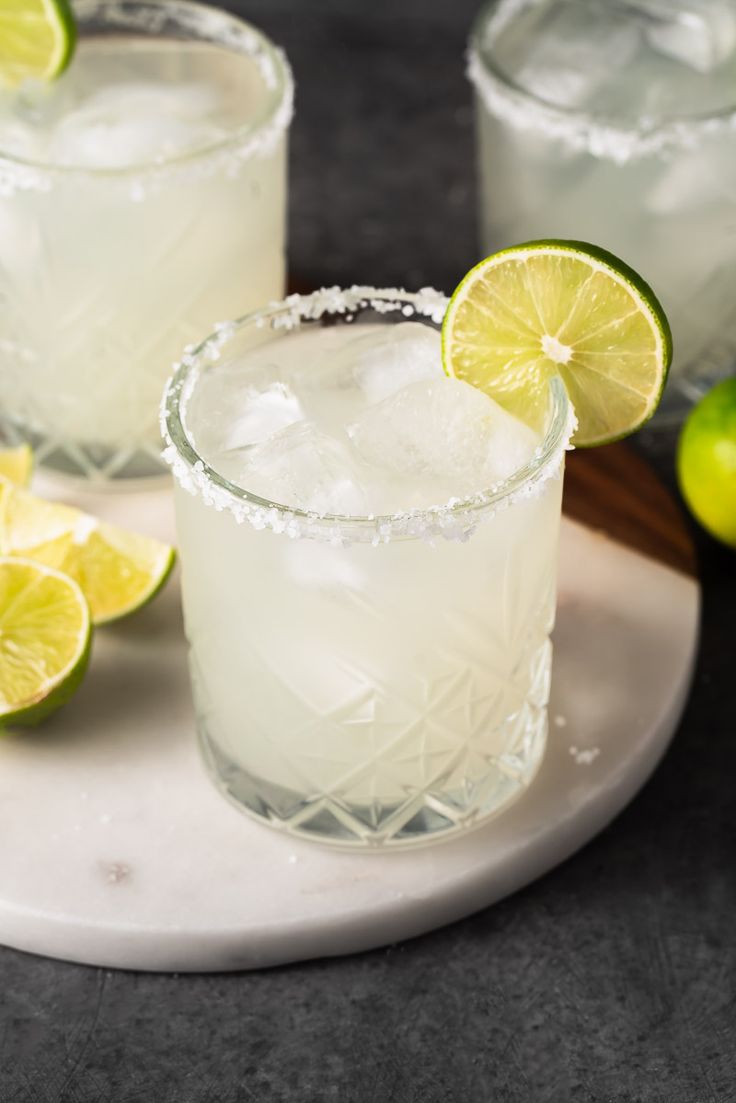 two glasses filled with lemonade and limes on a white plate next to some limes