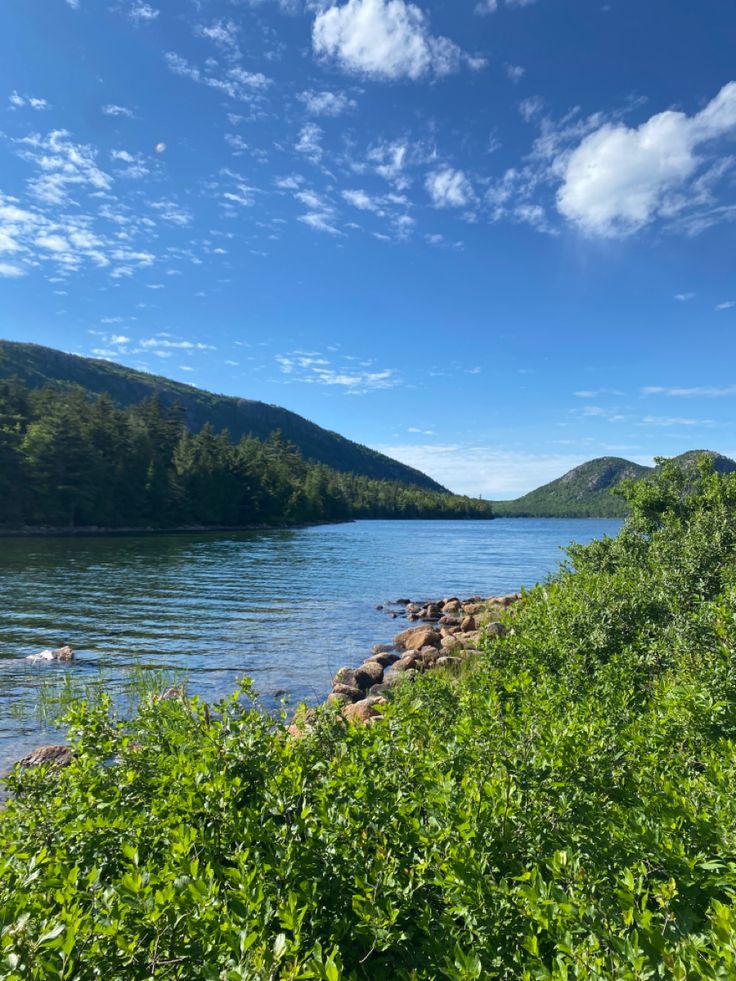 a lake surrounded by trees and rocks under a blue sky with wispy clouds