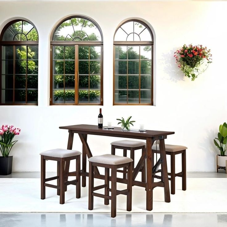 a dining table with four stools in front of three windows and potted plants