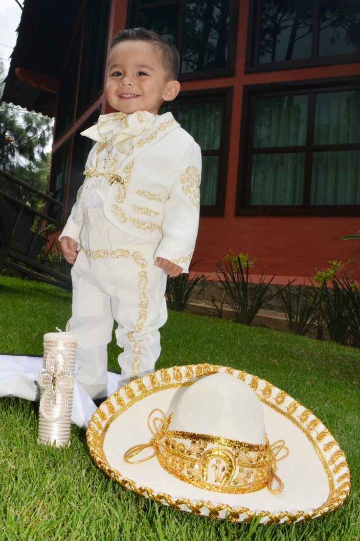 a little boy dressed in white and gold standing next to a hat on the grass