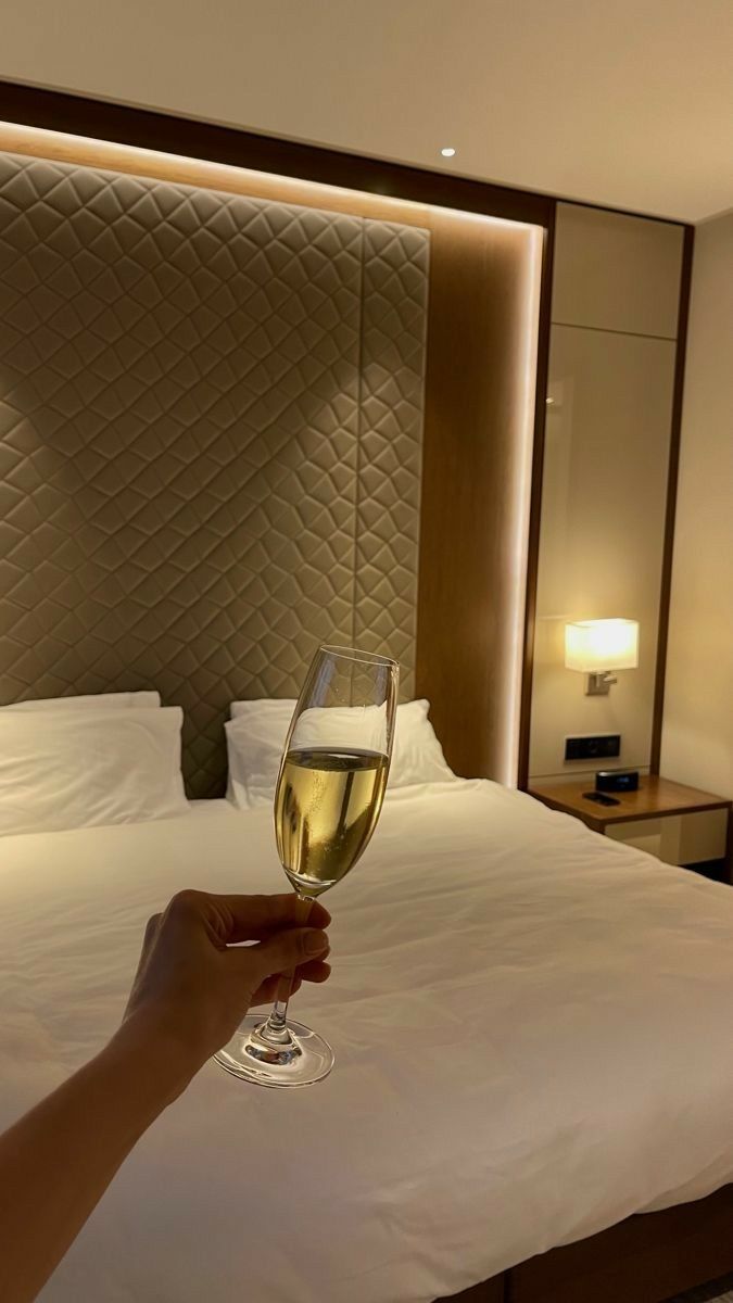 a person holding a glass of wine in front of a bed