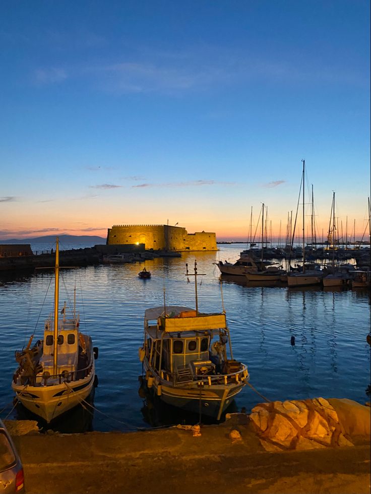 several boats are docked in the water at dusk near a large building with a castle on it's side