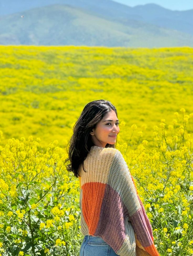 a woman standing in the middle of a field with yellow flowers and mountains behind her