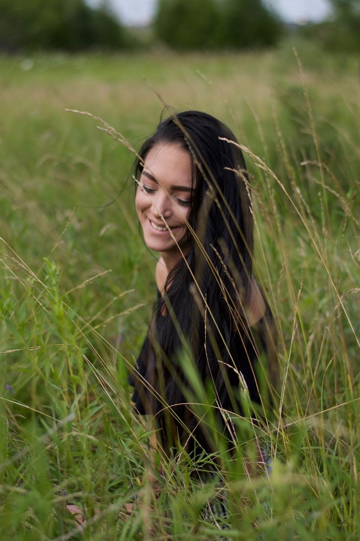 a woman is sitting in tall grass and smiling at the camera with her eyes closed