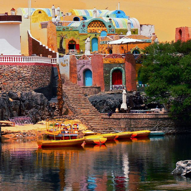 colorful buildings are reflected in the water near some rocks and trees, while another boat is on the riverbank