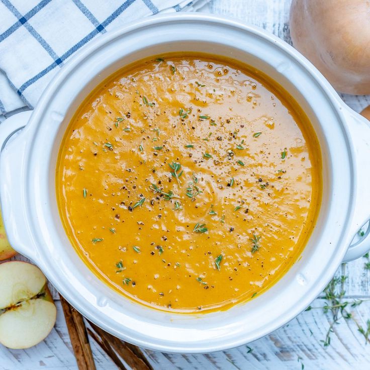 a white bowl filled with carrot soup next to an apple and cinnamon sticks on a table