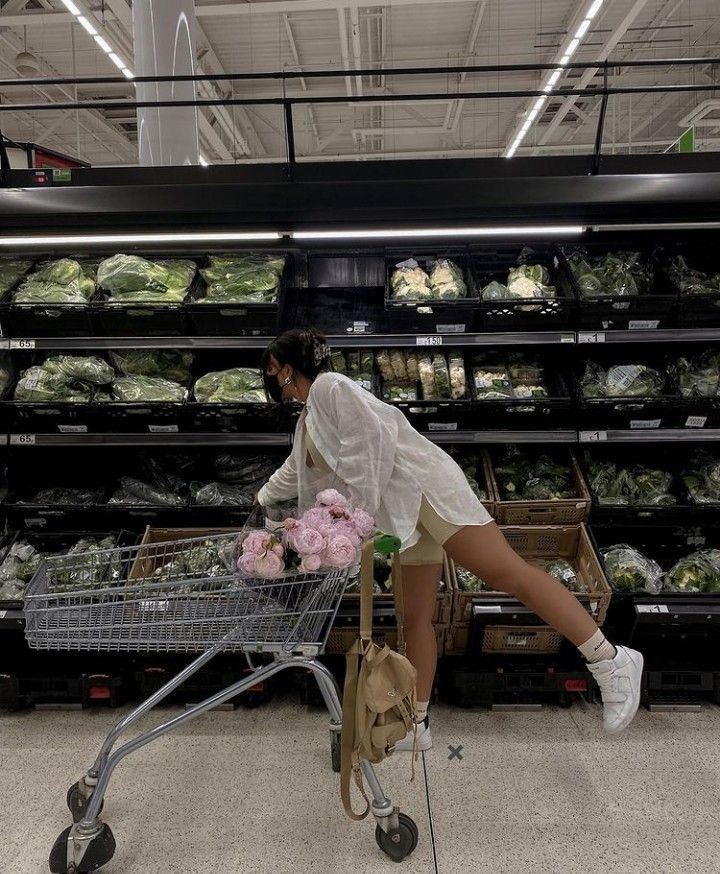 a woman pushing a shopping cart through a grocery store filled with vegetables and flowers,