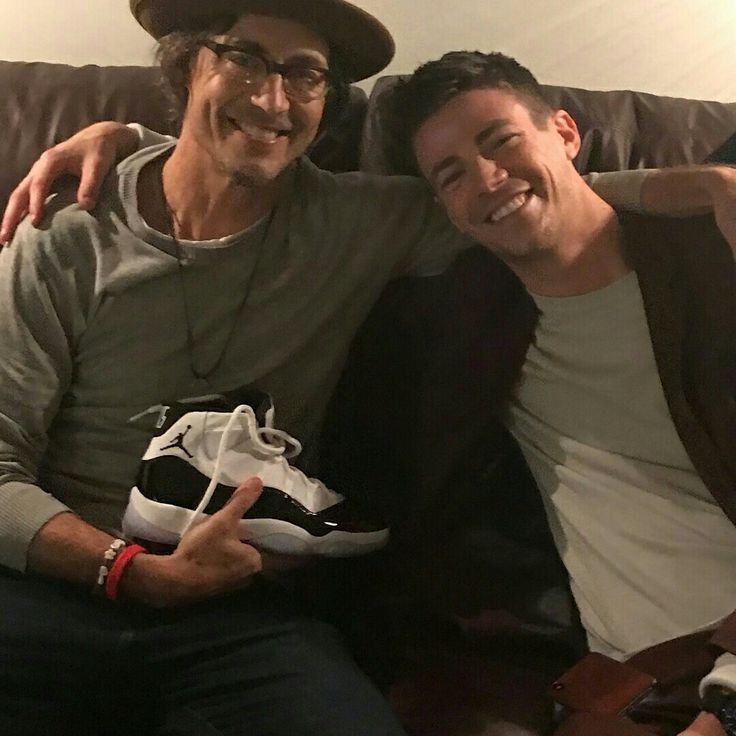 two men sitting on a couch with one holding a sneaker and the other smiling