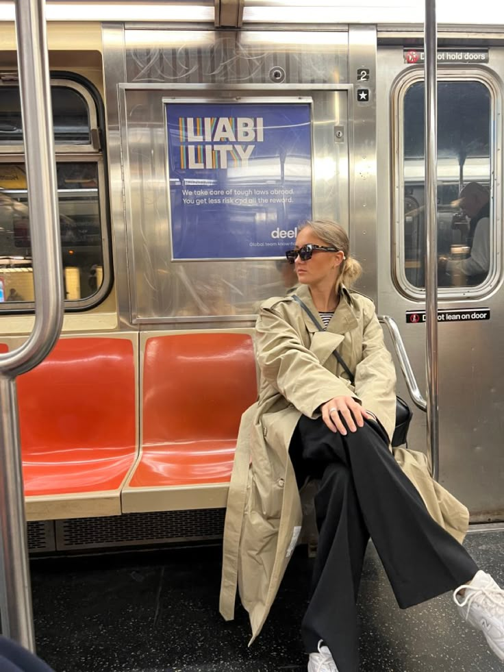 a woman sitting on a subway train with her legs crossed, wearing sunglasses and a trench coat