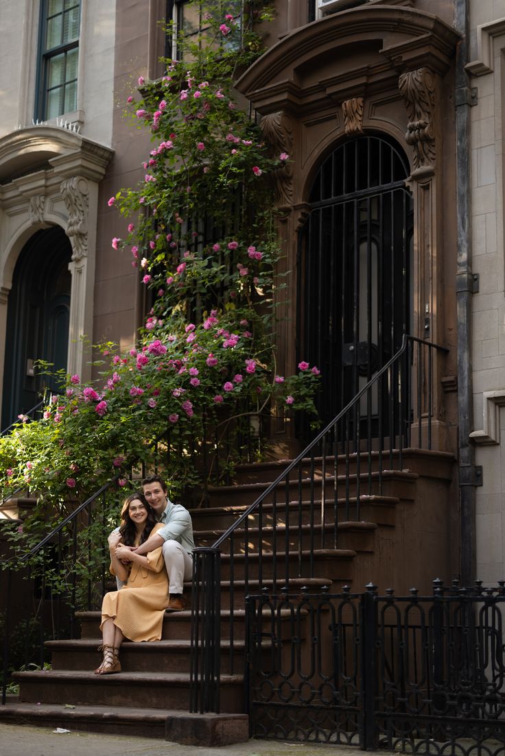 a man and woman sitting on the steps of a building in front of pink flowers