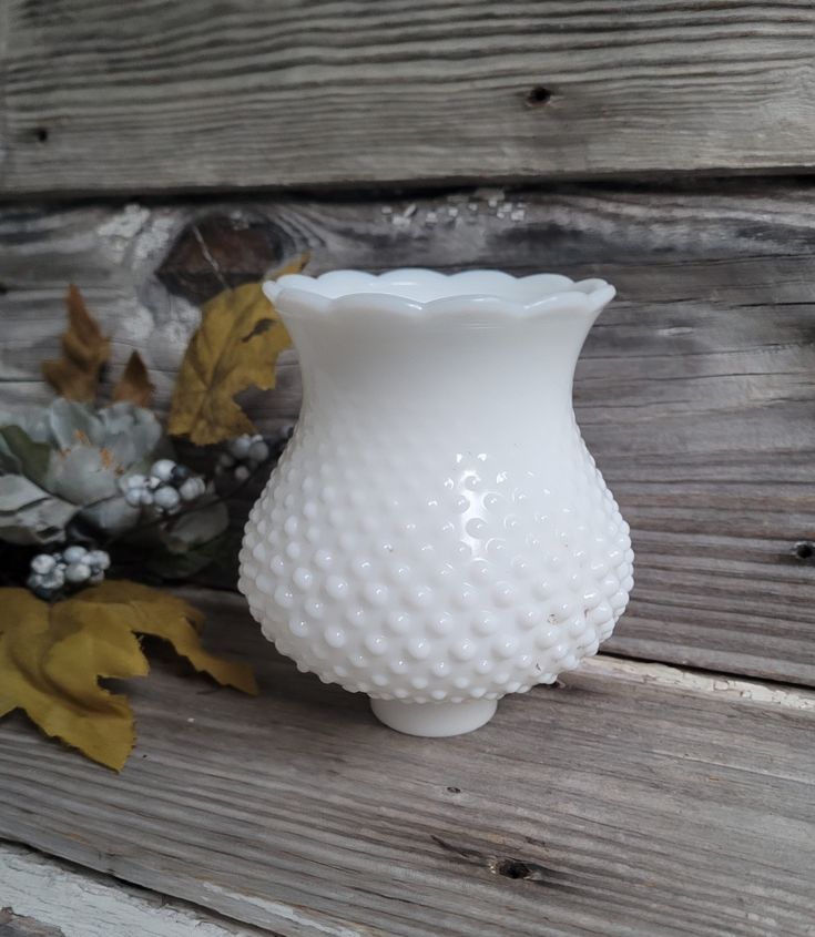 a white glass vase sitting on top of a wooden table next to leaves and flowers