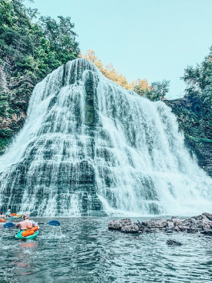 two people are kayaking in the water near a large waterfall that is surrounded by trees