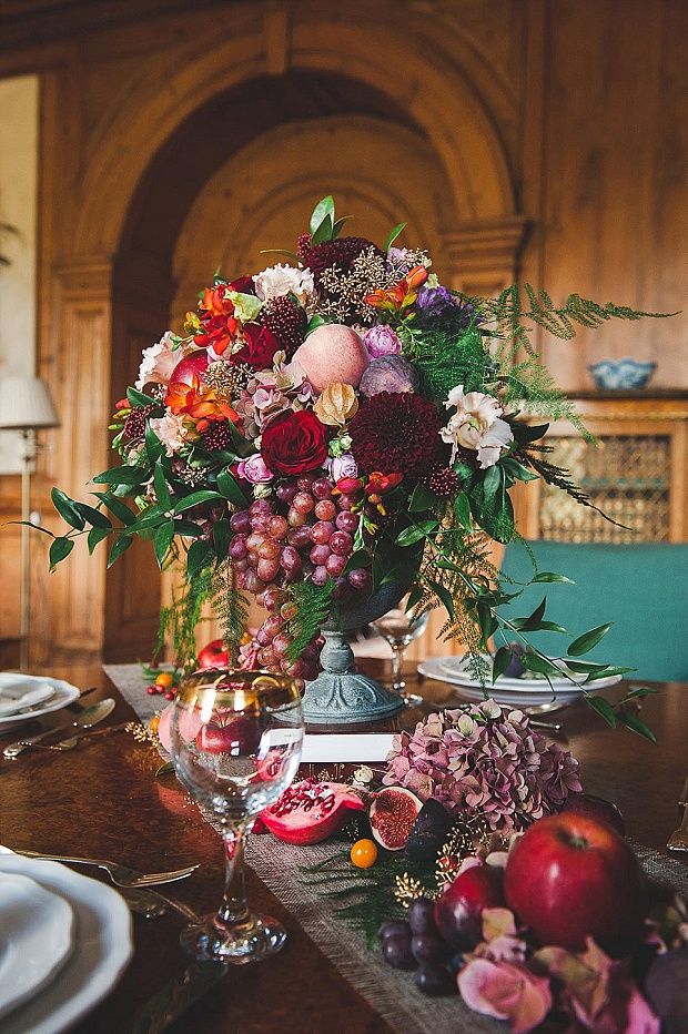 an arrangement of flowers and fruit on a table with wine glasses, plates and utensils