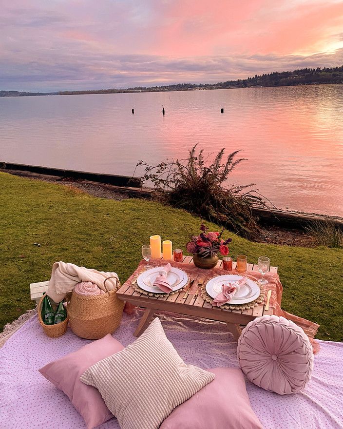 a picnic table set up on the grass by the water at sunset with candles, plates and napkins