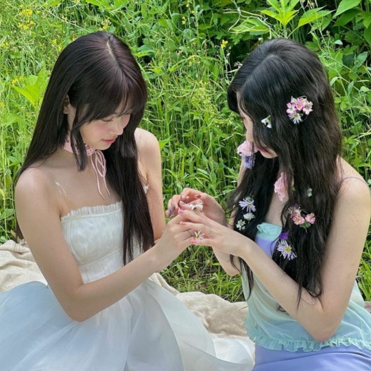 two young women sitting next to each other in front of green plants and flowers on the ground