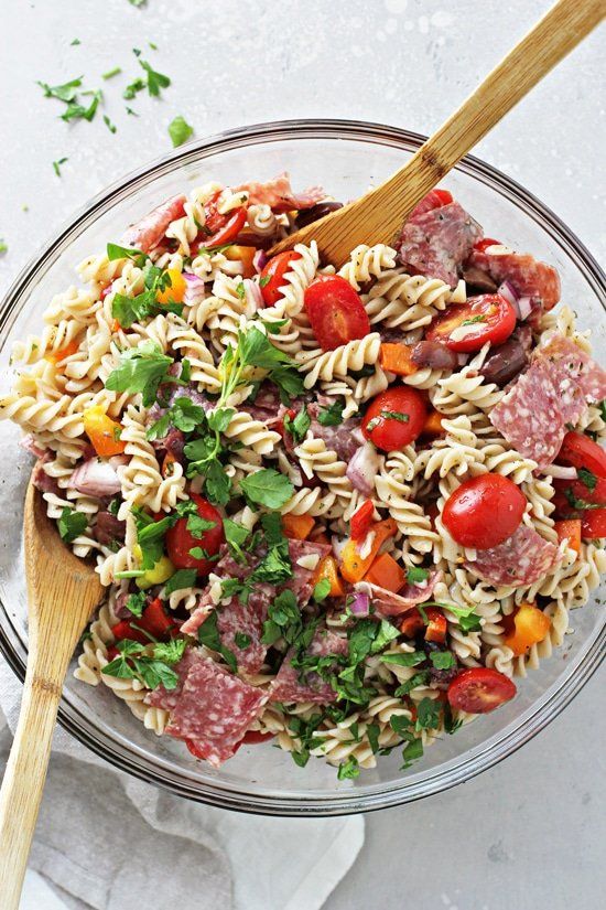 a glass bowl filled with pasta salad on top of a white tablecloth next to a wooden spoon