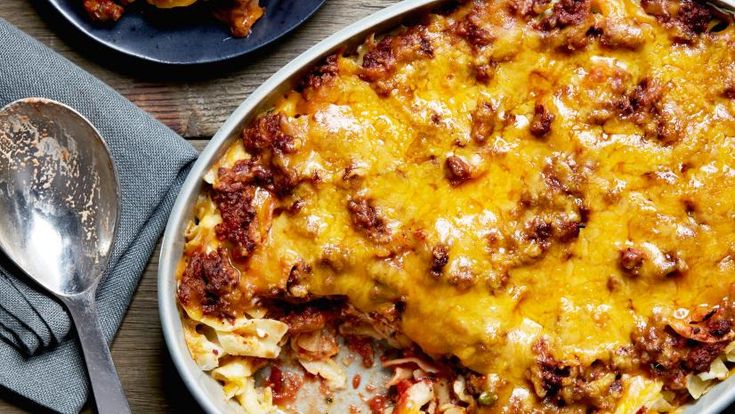 a casserole dish with meat and cheese in it on a wooden table next to two silver spoons