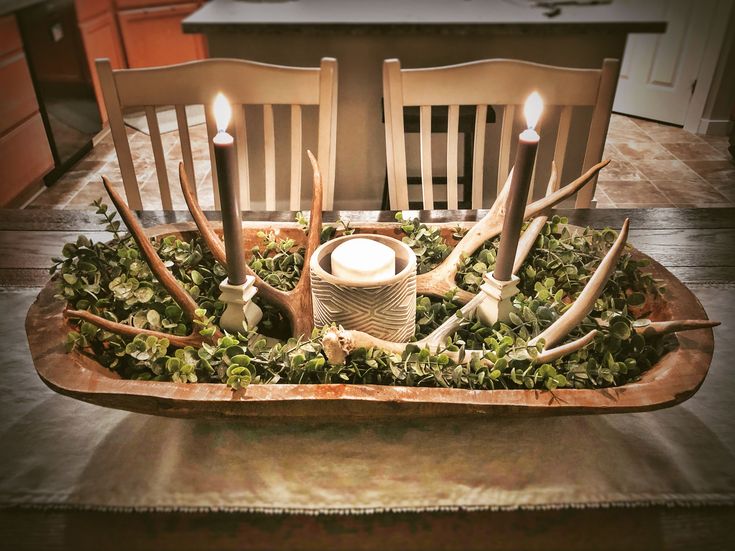 an arrangement of candles and antlers in a wooden tray with greenery on the table