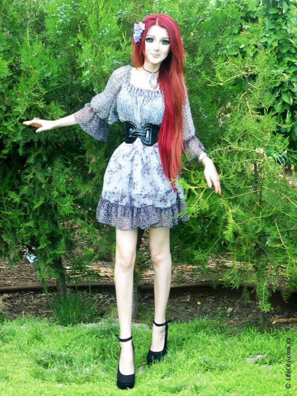 a doll with long red hair standing in the grass next to bushes and shrubbery