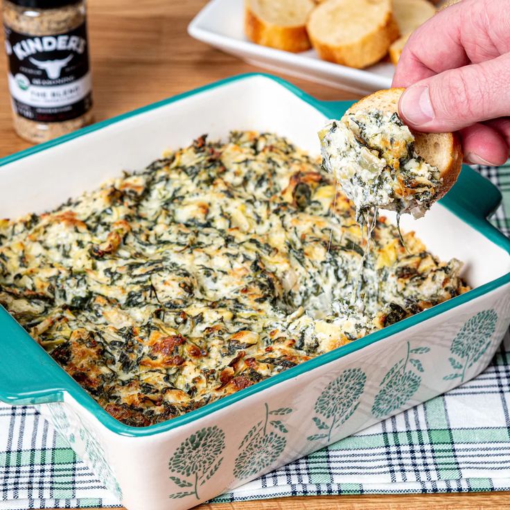 a hand holding a piece of bread over a casserole dish with spinach and cheese