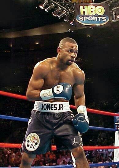 a man standing next to a boxing ring wearing black trunks and blue gloves with his hands on his hips