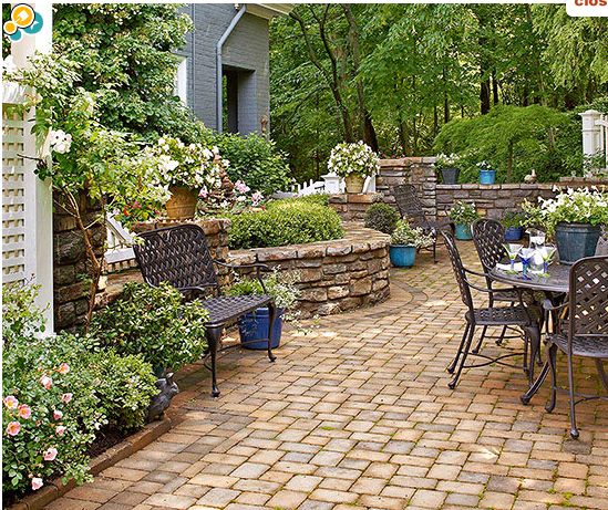 a brick patio with chairs and tables surrounded by flowers, shrubs and trees in the background