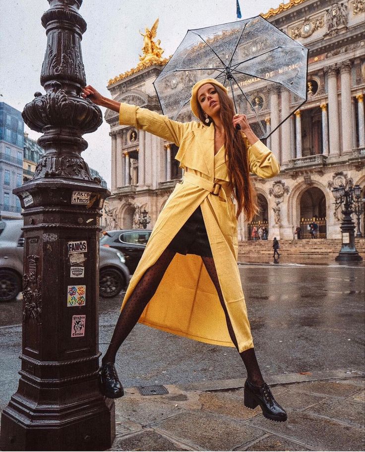 a woman in yellow coat and black pants holding an umbrella over her head while standing next to a lamp post