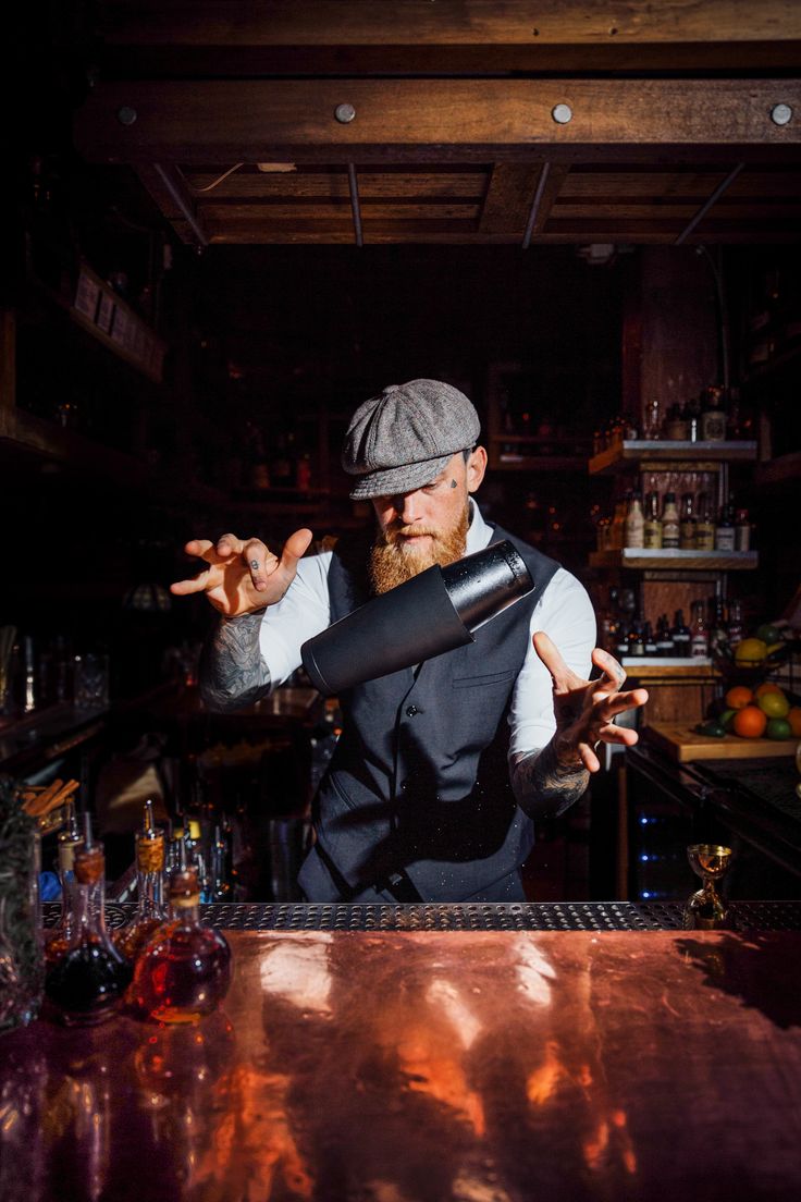 Bartender with beard and vintage hat flipping cocktail shaker mid air at the bar with bottles and wooden shelves around him Bartenders Photography, Boston Style, Most Popular Cocktails, Bartender Tools, Tiki Mugs, Popular Cocktails, Boston Fashion, Restaurant Photography, Cocktail Shaker Set