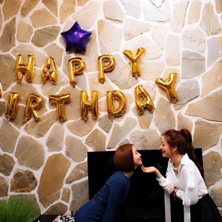 two women in front of a stone wall with the words happy birthday written on it