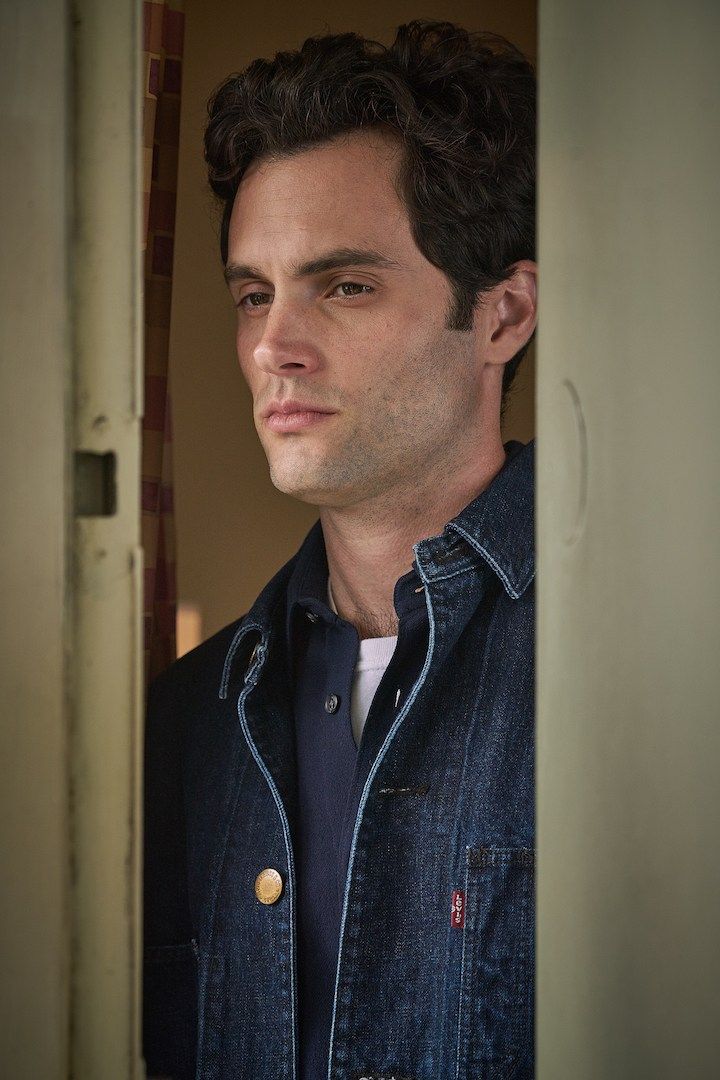 a man with curly hair wearing a denim jacket and looking into the distance while standing in front of a door