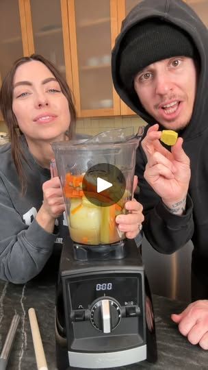 two people standing in front of a blender filled with fruit and vegetables, one holding a lemon