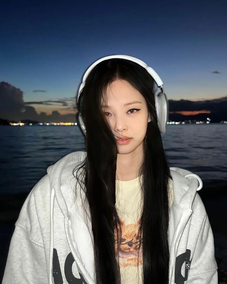 a girl with headphones on standing by the water at night, looking into the camera