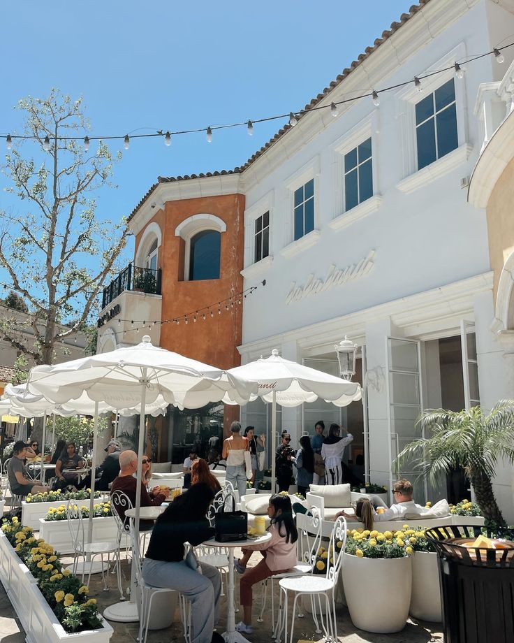 people are sitting at tables outside in front of a building with white umbrellas and potted plants