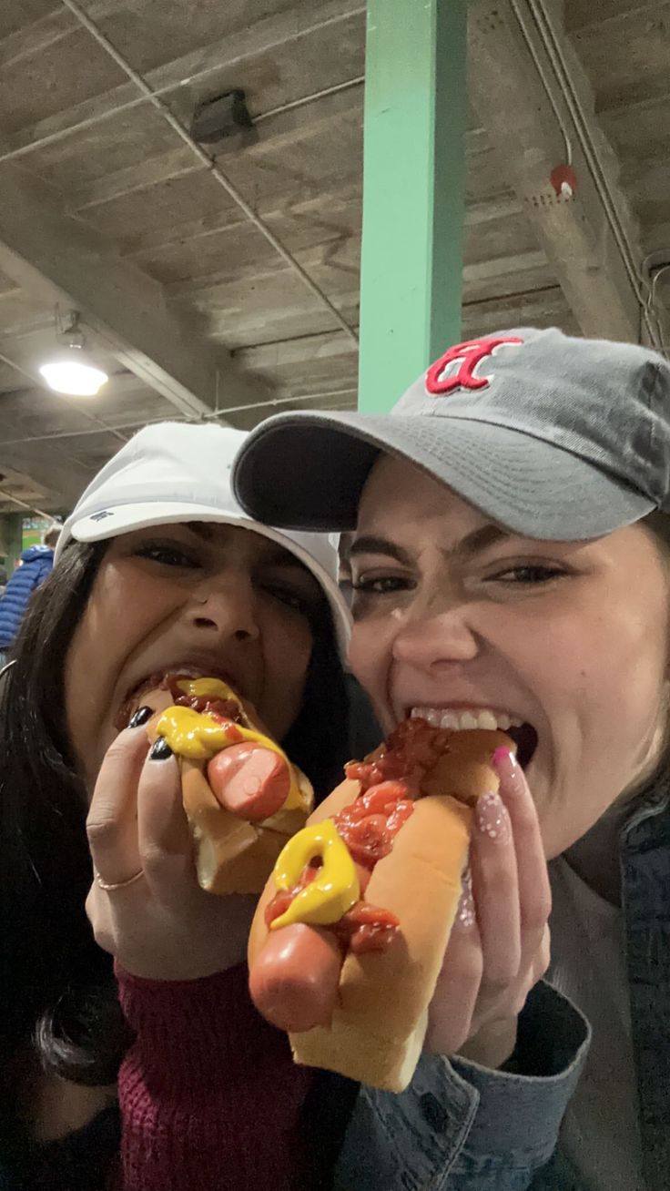 two women are eating hot dogs together in a subway station, one is wearing a hat and the other has a baseball cap