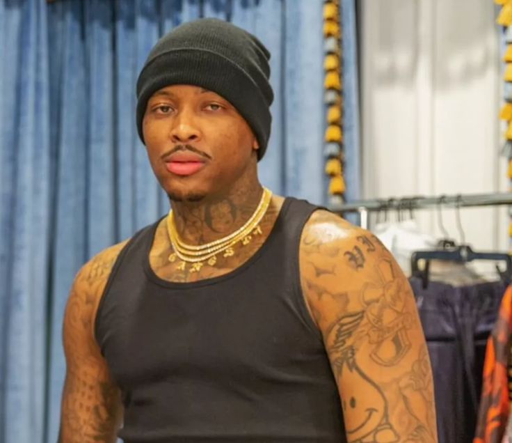 a man in a black tank top with tattoos on his arms and chest standing next to a blue curtain