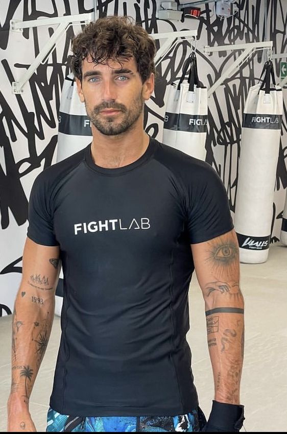 a man standing in front of a wall with graffiti on it and wearing a black shirt that says fightlab