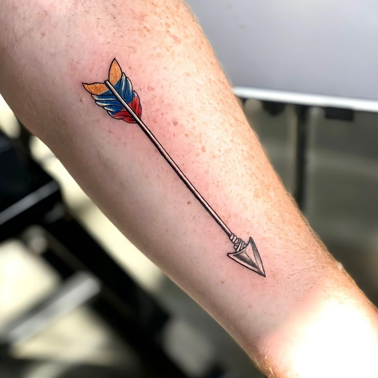 a small arrow tattoo on the arm with an orange and blue feather sitting on it