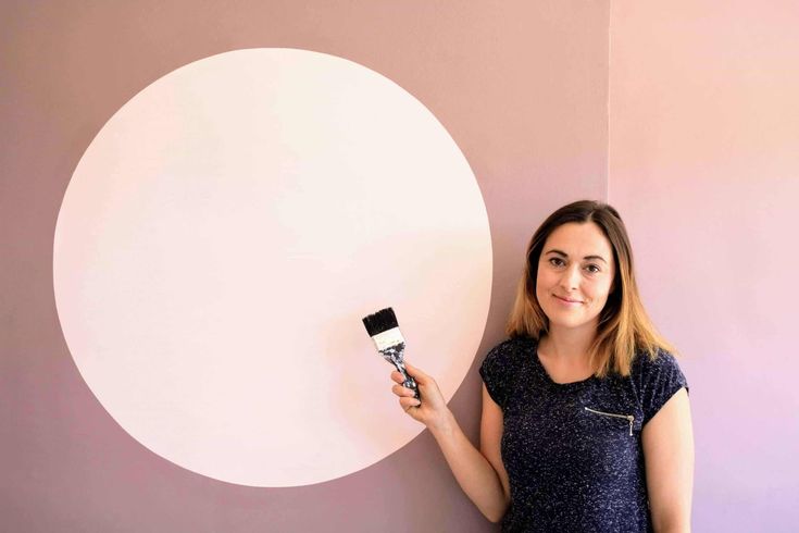 a woman standing next to a wall with a paintbrush in her hand and an oval painting on the wall behind her