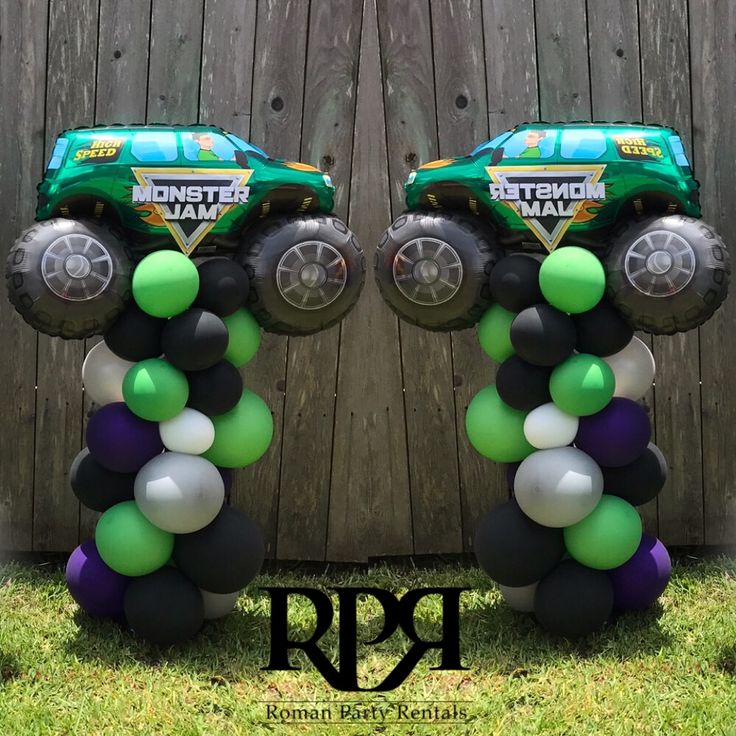 two monster trucks are decorated with balloons in front of a wooden fence, and one truck is on top of the balloon column