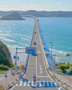 two people crossing the street on a long bridge over looking the ocean and hills in the distance